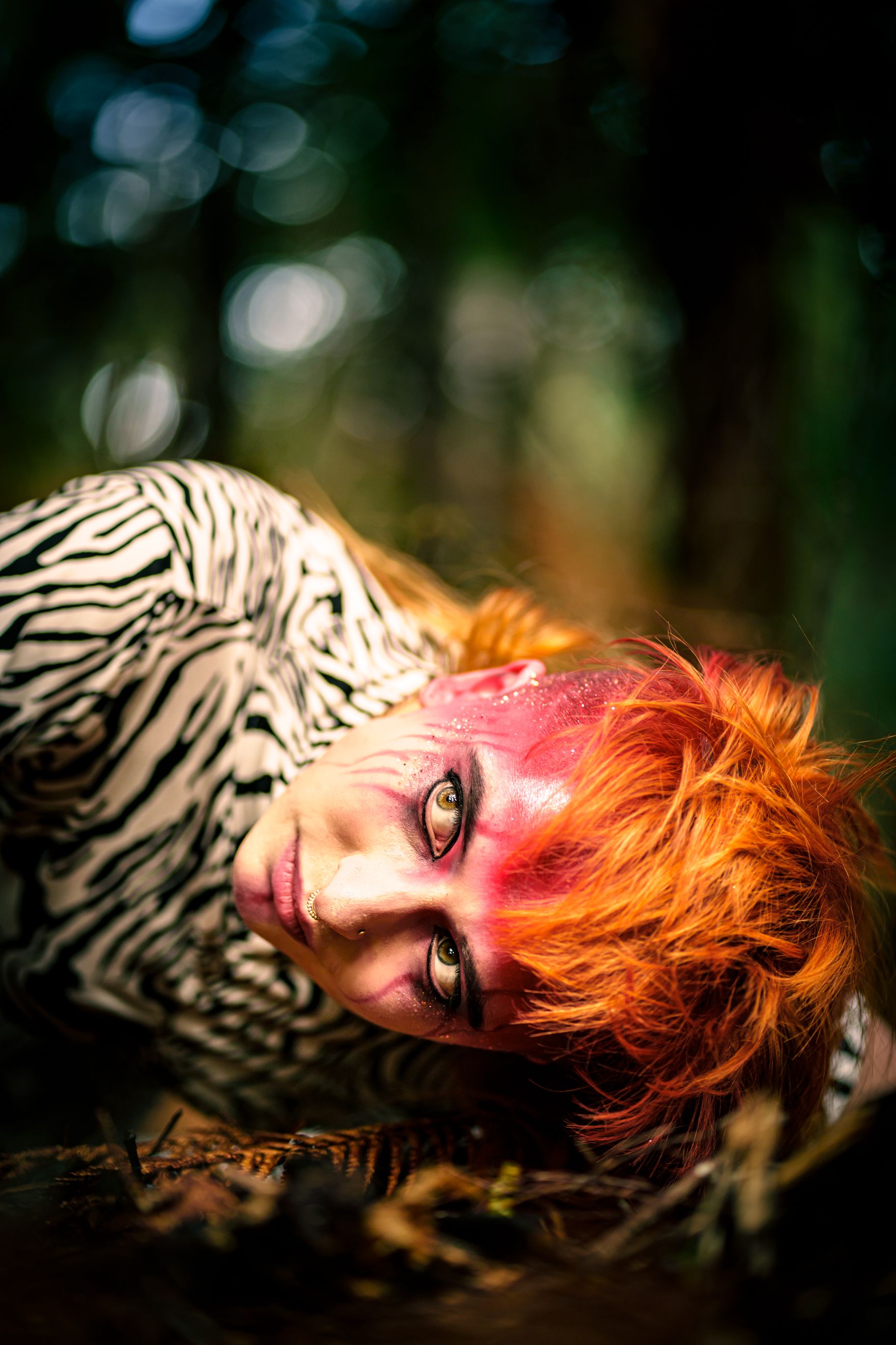 A close-up of a person, lying in their side and looking past the camera, they have bright orange hair and colourful facepaint